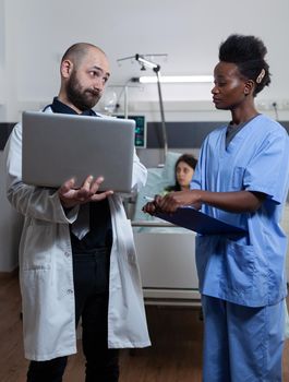 Doctor holding laptop unhappy with patient lab results looking with disapproval at nurse holding clipboard in hospital ward. Physician discussing medical treatment with health care worker.