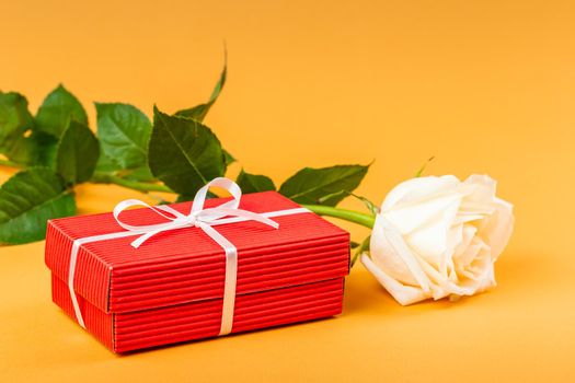 Red gift box with a white ribbon and white rose flower over vibrant orange background. Valentines Day or Birthday gift concept 