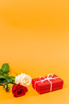 Red gift box with a white ribbon and two red and white roses over vibrant orange background. Vertical composition with space for text. Valentines Day or Anniversary gift concept. 