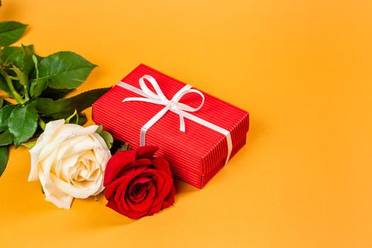 Red gift box with a white ribbon and two red and white roses over vibrant orange background. Valentines Day or Birthday gift concept 