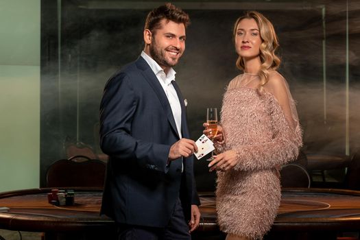 Couple of successful poker players standing near gaming table with stack of betting chips on background of smoky interior of empty casino hall. Confident woman champagne, smiling man showing two aces