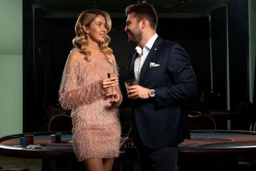 Smiling young attractive blonde woman and stylish bearded man standing near card game table with stacks of chips in casino, holding glasses with drinks. Successful poker players celebrating win