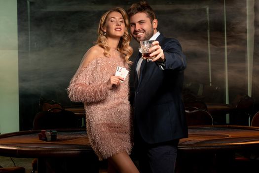 Portrait of confident stylish young female poker player standing in casino near gaming table holding pair of aces and smiling bearded man raising glass of whisky with view of winner. Gambling success