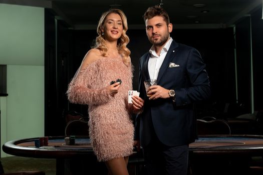 Portrait of stylish elegant blonde girl and bearded man happy with successful poker game standing with winning set of two aces, chips and glass of drink near gaming table in casino