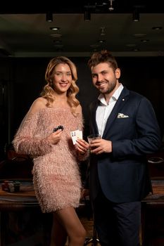 Smiling bearded guy with glass of drink and elegant young blonde holding pair of ace and betting chips standing near gaming table in casino, celebrating winning poker. Successful gambling concept