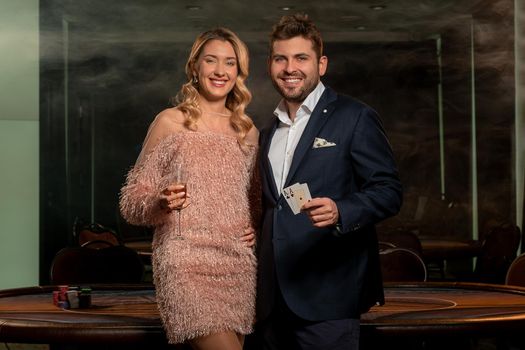 Happy young elegant couple posing in casino near poker table. Smiling woman holding glass of champagne, cheerful man showing winning card set of pair of aces. Gambling concept