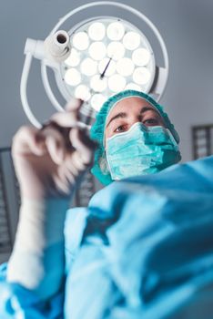 From below woman surgeon in medical uniform using professional tools while standing under bright light in operating theater. High quality photo