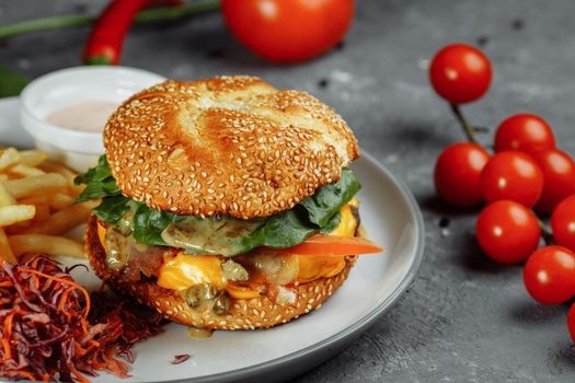 Turkey burger on a bun with cheese, lettuce, tomatoes and fresh cucumber. Burger with cutlet of turkey meat on a gray background with french fries sauce and salad.