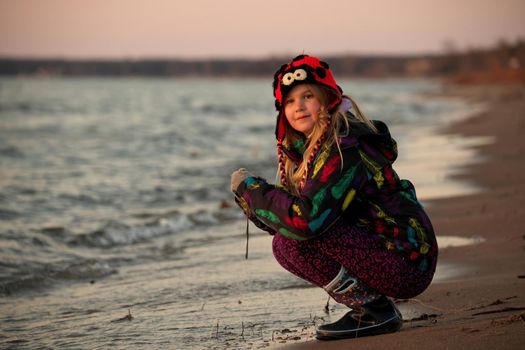A 9 year old budding photographer practices her camera skills at sunset at the beach. It is cold and she wears a winter jacket and ladybug toque. High quality photo