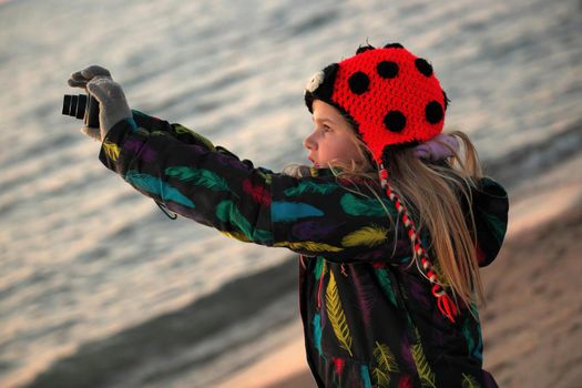 A 9 year old budding photographer practices her camera skills at sunset at the beach. It is cold and she wears a winter jacket and ladybug toque. High quality photo