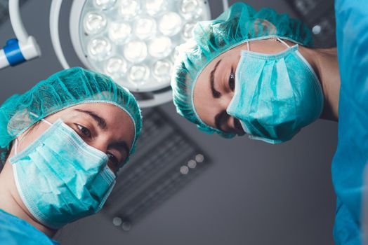 From below female surgeons in medical uniform using professional tools while standing under bright light in operating theater. High quality photo
