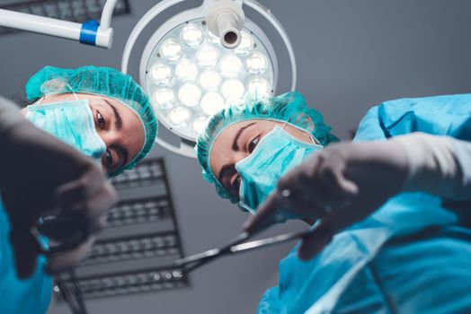From below female surgeons in medical uniform using professional tools while standing under bright light in operating theater. High quality photo