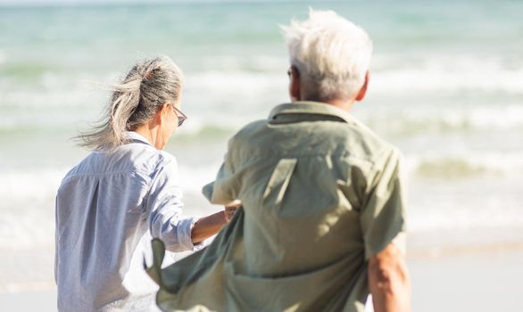 Happy Asian senior man and woman couple holding hands walking to the beach sunny with bright blue sky, Romantic elderly enjoy Travel summer vacation, plan life insurance at retirement couple concept