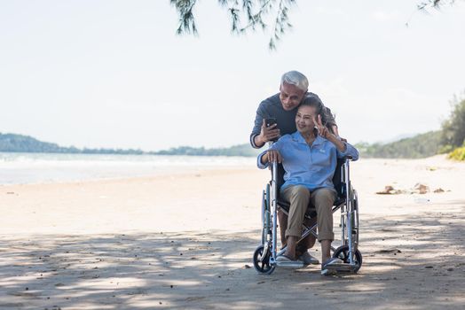 Happy Asian elderly woman sitting in wheelchair and husband is a wheelchair user smartphone taking selfie on the beach, summer vacation, Retirement couple concept