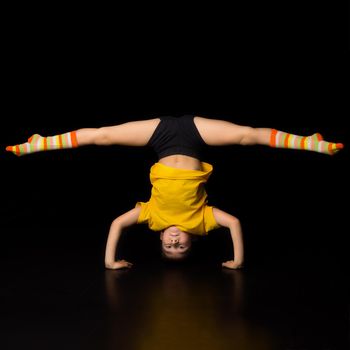Young boy doing handstand and splits acrobatic element. Boy gymnast or dancer wearing sportswear and striped socks performing gymnastic exercise against isolated black background in studio