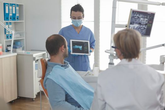 Dentist assistant wearing face mask showing teeth radiography using tablet to patient discussing medical procedure during stomatology examination in dental office. Sick man having oral infection