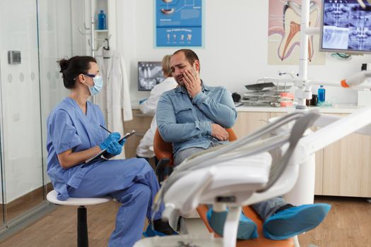 Patient with toothache sitting on dental chair during medical consultation in stomatological office room. Orthodontist nurse woman explaining oral hygiene writing healthcare treatment on clipboard