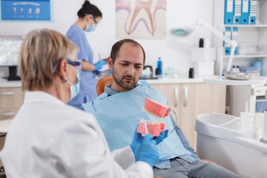 Senior woman doctor showing artificial jaw model to patient with toothache discussing carier treatment during stomatology consultation in dentistry office room. Stomatologist explaining oral hygiene