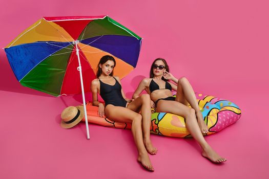 Two young female friends in swimsuits sitting on inflatable mattress under colored sun umbrella on pink studio background, enjoying imaginary seaside vacation. Summer beach holiday waiting concept