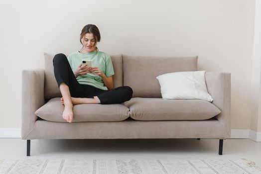 Image of happy optimistic young woman sitting indoors at home and shopping or social media using mobile phone on sofa at home