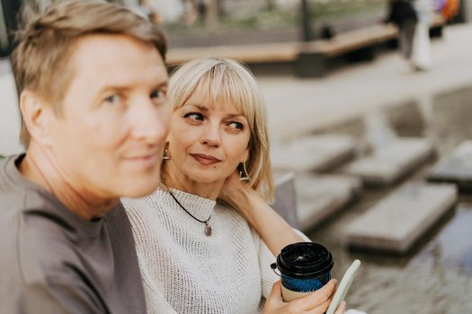 An adult mature happy couple in love sitting on bench outdoors in city street park. A blonde caucasian man and woman spend time together and drinking coffee. Senior wife and husband walking outside