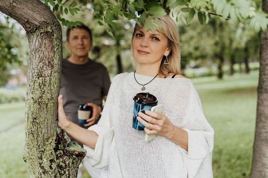 An adult mature happy couple in love hugging outdoors in city park. A blonde caucasian man and woman spend time together and drinking coffee. Senior wife and husband walking outside