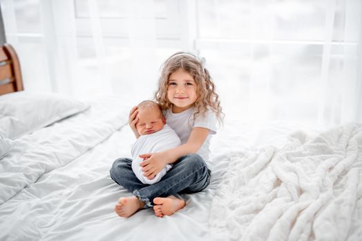 Cute little curly hair girl sitting on the bed and holding adorable newborn baby. Beautiful portrait of sister and her sibling infant child together at home. Happy family moments with children