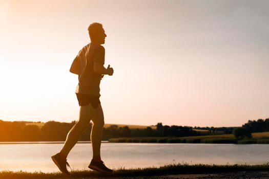 Young man doing cardio workout, exercising and running early in the morning at dawn near a beautiful lake. The athlete is preparing for the marathon.