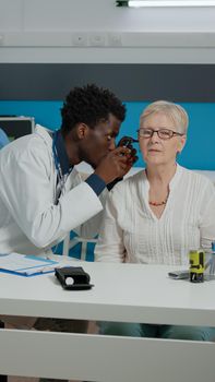 Man doctor using otoscope for ear examination of senior patient sitting at desk. Young otologist with professional tool in medical cabinet doing consultation for old woman with disease