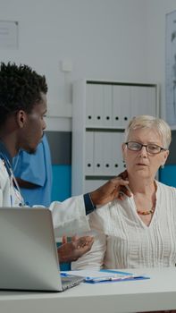 Young medic consulting sick elderly woman with disease in medical office. Doctor doing healthcare checkup on old patient at desk while nurse and senior man on bed talking in background