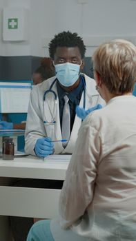 Young medic wearing face mask while consulting elder patient behind plexiglass wall at medical facility. Doctor helping senior woman with healing treatment for healthcare during pandemic