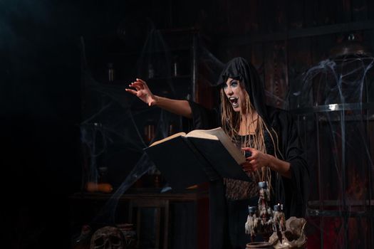 Halloween concept. Witch dressed black hood with dreadlocks standing dark dungeon room use magic book for conjuring magic spell. Female necromancer wizard gothic interior put her hand forward scream