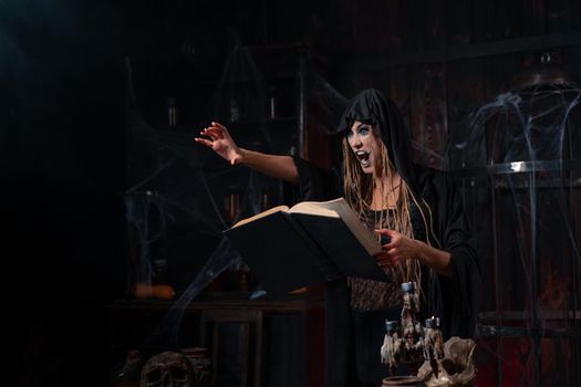 Halloween concept. Witch dressed black hood with dreadlocks standing dark dungeon room use magic book for conjuring magic spell. Female necromancer wizard gothic interior put her hand forward scream