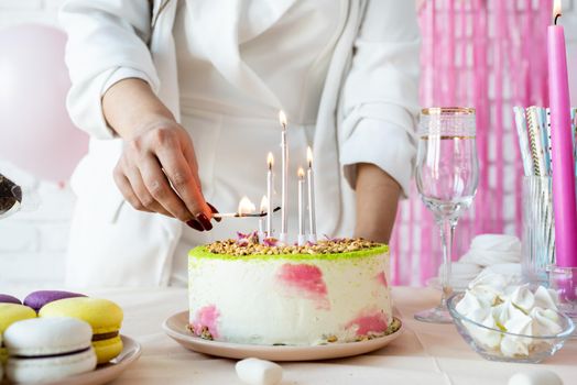 Birthday party. Birthday tables. Attractive woman in white party clothes preparing birthday table with cakes, cakepops, macarons and other sweets, lighting the candles