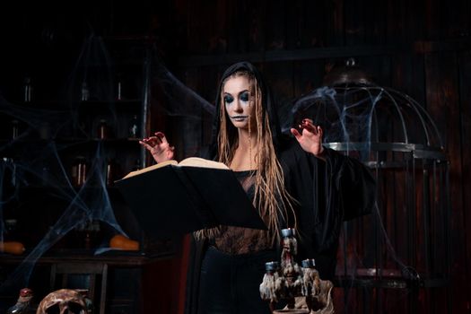 Halloween concept. Witch dressed black hood standing dark dungeon room use magic book for conjuring magic spell. Female necromancer wizard gothic interior with cage, spider web