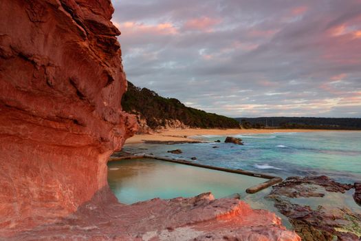 Aslings beach rock pool is built into the cliffs of which dramatic red rock is a feature. Peach and pink in the clouds at sunrise. Beautiful landscape and ocean baths New South Wales Australia