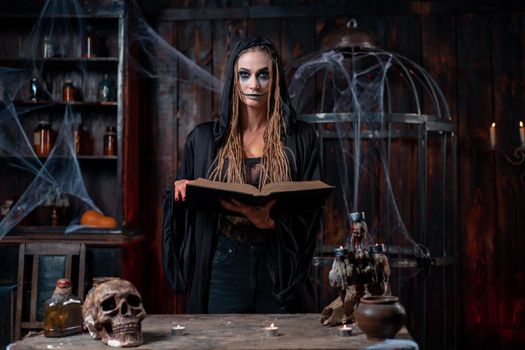 Halloween concept. Witch dressed black hood standing dark spooky room use magic book for conjuring magic spell. Female necromancer wizard gothic interior with skull, cage, spider web