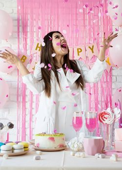 Birthday party. Birthday tables. Attractive brunette woman in white party clothes preparing birthday table with cakes, cakepops, macarons and other sweets, throwing pink confetti