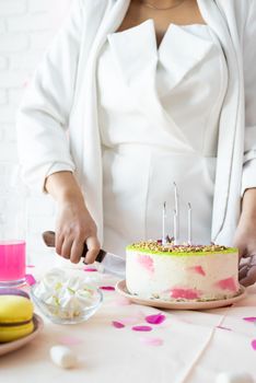Birthday party. Birthday tables. Attractive woman in white party clothes preparing birthday table with cakes, cakepops, macarons and other sweets, cutting a cake