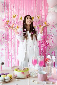 Birthday party. Birthday tables. Attractive brunette woman in white party clothes preparing birthday table with cakes, cakepops, macarons and other sweets, throwing pink confetti