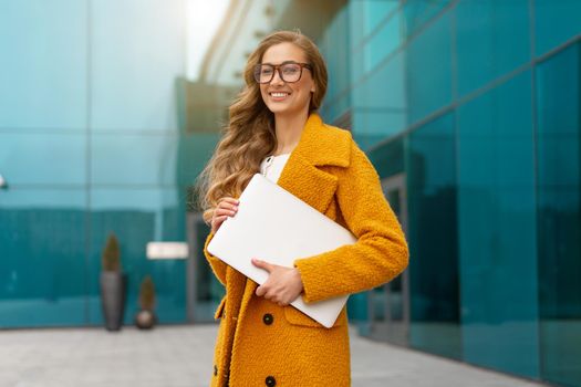 Business woman with laptop dressed yellow coat standing outdoors corporative building background Caucasian female business person eyeglasses on city street near office building Stylish businesswoman