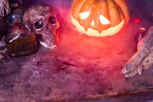 Halloween. Scary Halloween pumpkin with carved face on table in dark room with human skull and animal skull. Copy space for text. Spooky atmosphere.