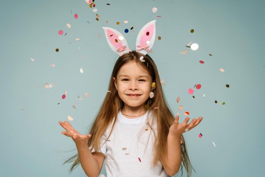 Beautiful happy little girl with bunny ears with confetti on a blue background celebrates easter