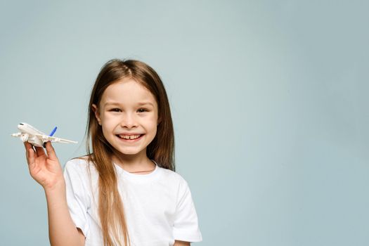 Little girl smiling while holding a fake airplane on a blue background and looking at the camera. Tourism and travel concept. Copy space.