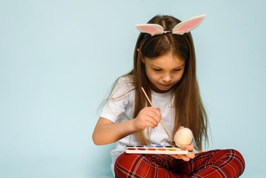 A little girl in rabbit ears paints eggs for Easter on a blue background in the studio