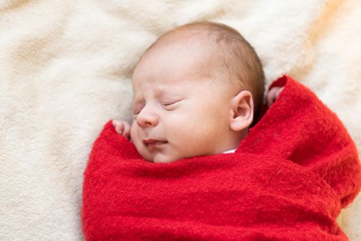 Top View Portrait First Days Of Life Newborn Cute Sleeping Baby In Santa Hat Wrapped In Red Diaper rug At White Garland Background. Merry Christmas, Happy New Year, Infant, Childhood, Winter Concept.