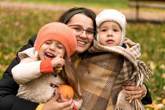 Young Woman Mom And Little Cute Preschool Minor Children In Orange Plaid At Yellow Fallen Leaves Nice Smiling Look At Camera In Cold Weather In Fall Park. Childhood, Family, Motherhood, Autumn Concept.