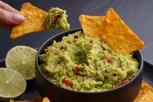 Closeup of woman hand with tortilla chips or nachos with fresh tasty guacamole dip in black plate on black background. High quality photo
