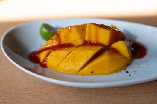 Close-up of half a lime and chopped mango with chili powder and Mexican chamoy sauce on white plate. Fresh tropical fruit slices with spicy and sour condiment on table. Healthy food and snacks