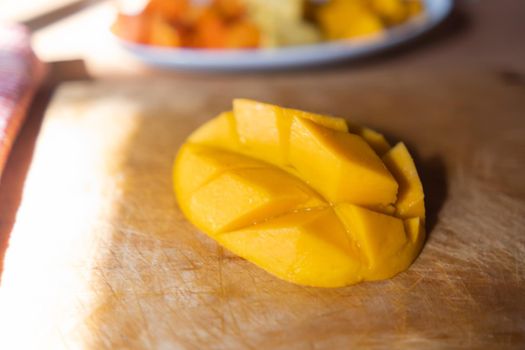 Close-up of diced mango on scratched wooden cutting board. Fresh yellow tropical fruit slice above wooden surface with blurry background. Healthy snack preparation